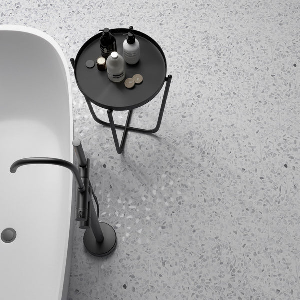 A modern bathroom with a freestanding white bathtub, a sleek silver faucet, and a round black side table holding various toiletries on a light terrazzo floor.