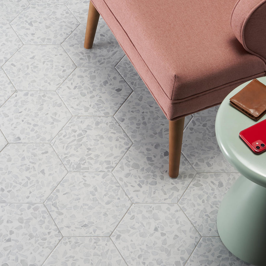 A pink upholstered chair next to a round green table with a red smartphone and a brown wallet on it, set on a white hexagonal tile floor with subtle gray patterns.