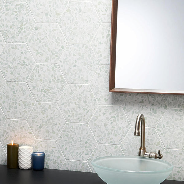 A bathroom countertop with three lit candles, a glass vessel sink, a silver faucet, and a wall covered in hexagonal tiles with a mosaic pattern. Part of a wooden-framed mirror is visible.