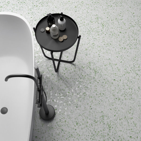 A modern bathroom with a white bathtub, a black side table holding bottles and jars, and terrazzo flooring with white and green speckles.