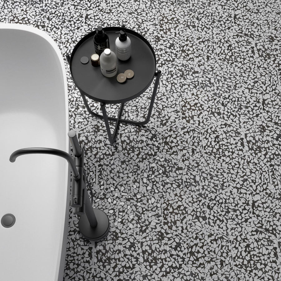 A white freestanding bathtub with a modern silver faucet stands beside a round black side table holding toiletries, set on a white and gray terrazzo-patterned floor.