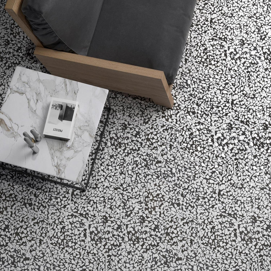 A marble coffee table with a magazine and decorative object sits next to a wooden sofa with a gray cushion, on a black and white terrazzo-patterned floor.