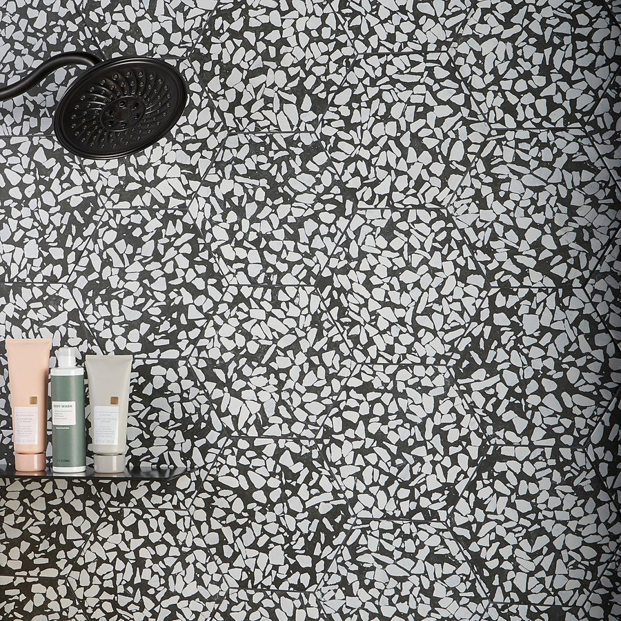 A black rain shower head and a shelf with bottles of toiletries are mounted on a wall with a black and white mosaic terrazzo tile pattern.