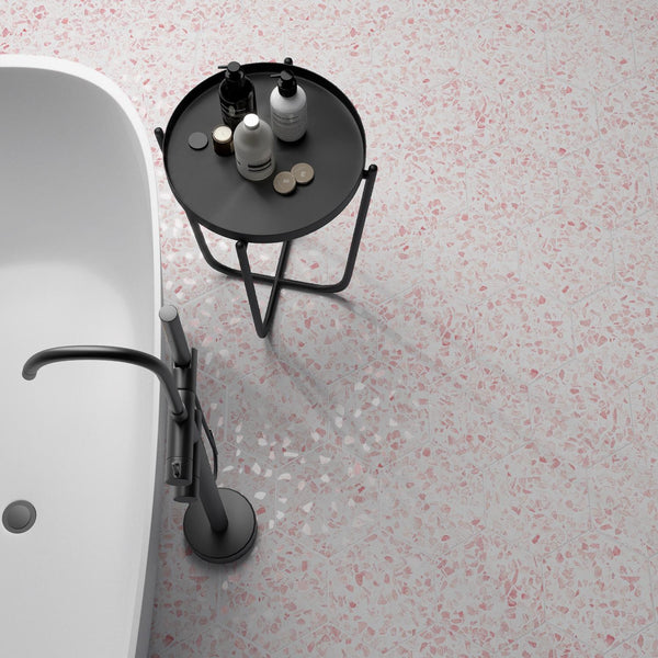A modern bathroom with a white bathtub, a sleek silver faucet, and a small round black table holding bottles and jars, all set on a light pink terrazzo floor.