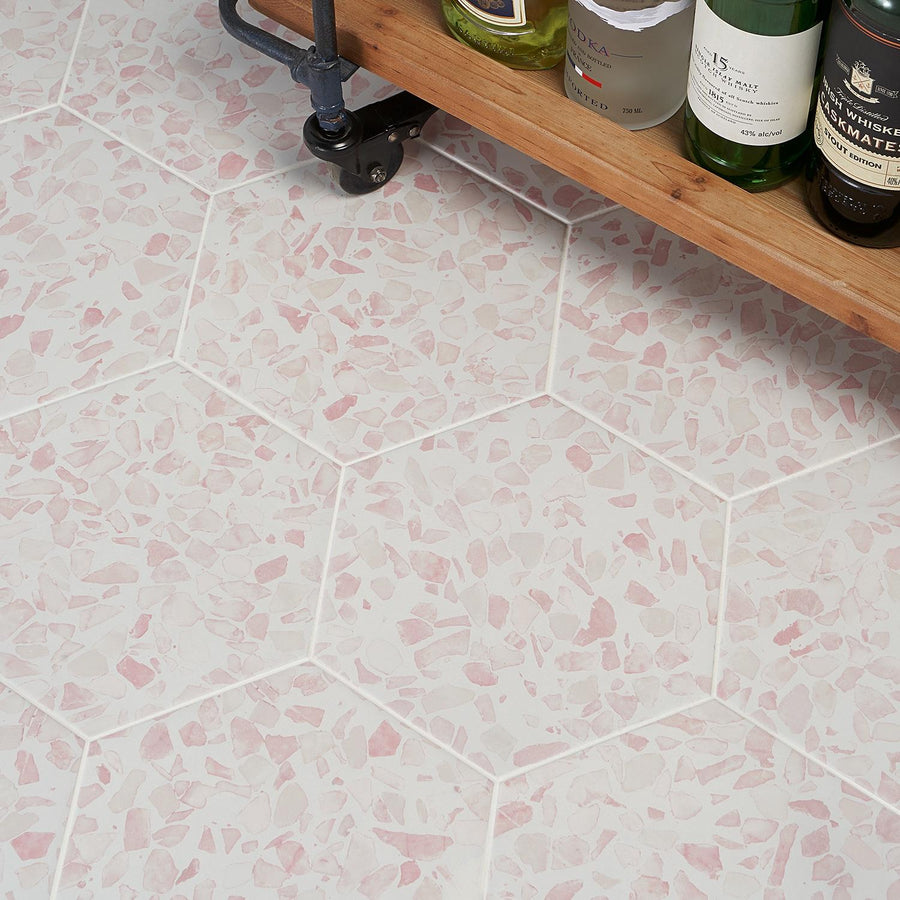 A wheeled cart with bottles of various alcoholic beverages stands on a white hexagonal tile floor with pink terrazzo-style patterns.