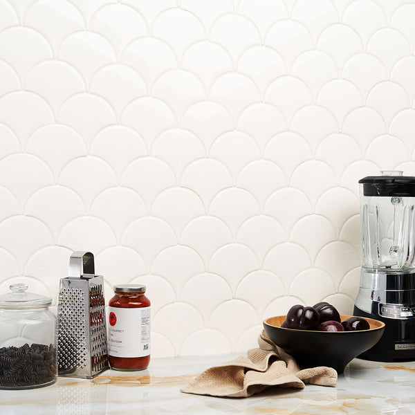 A kitchen counter with a glass jar of cookies, a grater, a jar of sauce, a bowl of plums, a beige cloth, and a blender, set against a white, scallop-patterned tile backsplash.