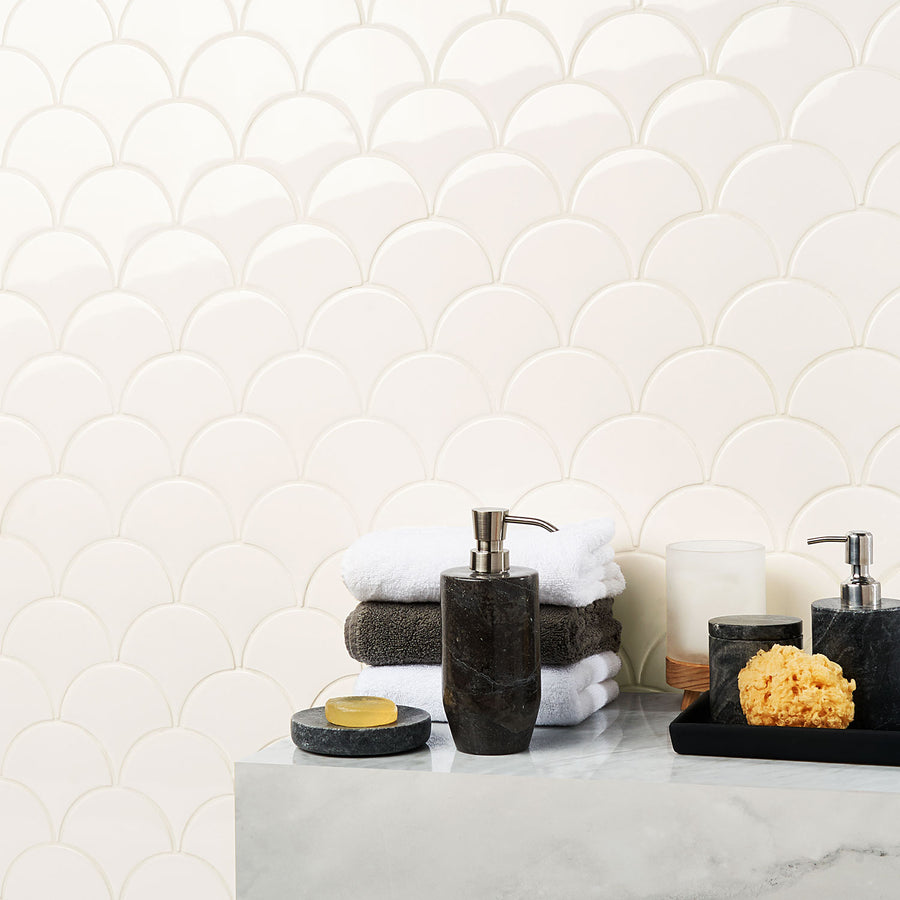 A bathroom countertop with stacked white and dark towels, a soap dispenser, a bar of soap on a dish, a natural sponge, a candle, and a tray, set against a white scallop-patterned tile wall.