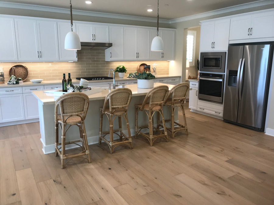 Modern kitchen with white cabinets, stainless steel appliances, light wood floors, an island with four wicker bar stools, pendant lights above, and decorative plants and bottles on the countertop.