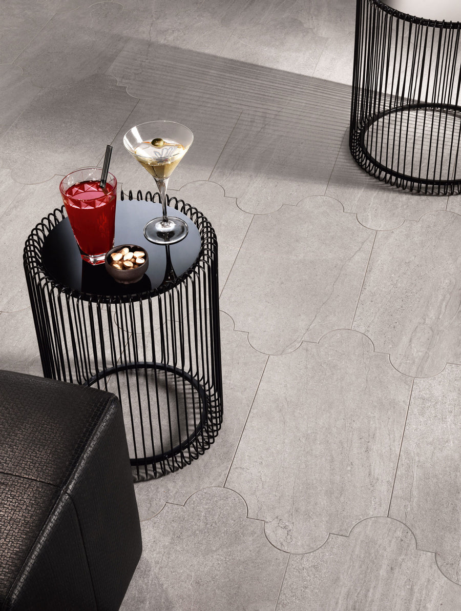 A small black round side table holds a red cocktail, a martini, and a bowl of snacks. The table has a wire frame design and sits on a light gray tiled floor beside a black cushioned seat.