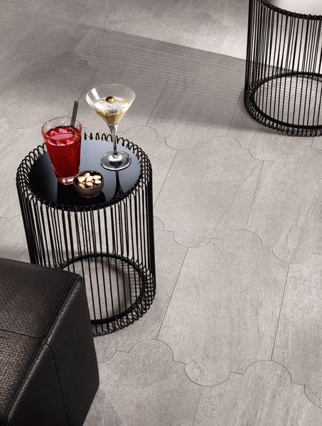 A small black round side table holds a red cocktail, a martini, and a bowl of snacks. The table has a wire frame design and sits on a light gray tiled floor beside a black cushioned seat.