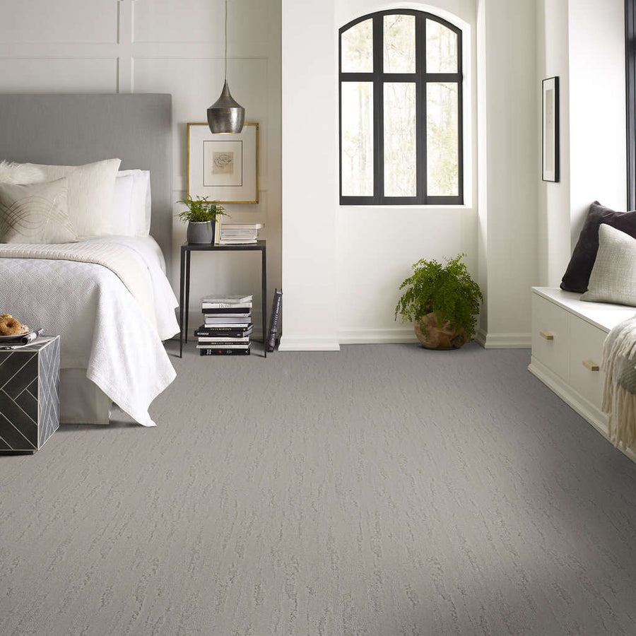 A modern bedroom with a gray carpet, white bedding, a nightstand with a lamp and plant, black window with natural light, and a built-in bench with a pillow and green plant. The room is bright and minimalistic.