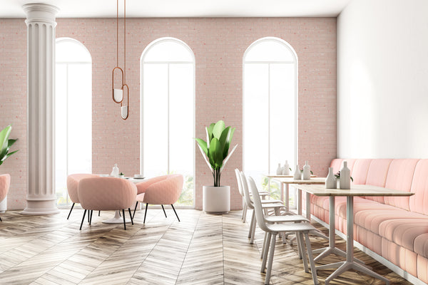 A bright, modern cafe with pink cushioned chairs and benches, round and rectangular tables, large arched windows, potted plants, and minimalist pendant lights, featuring light wood herringbone flooring.