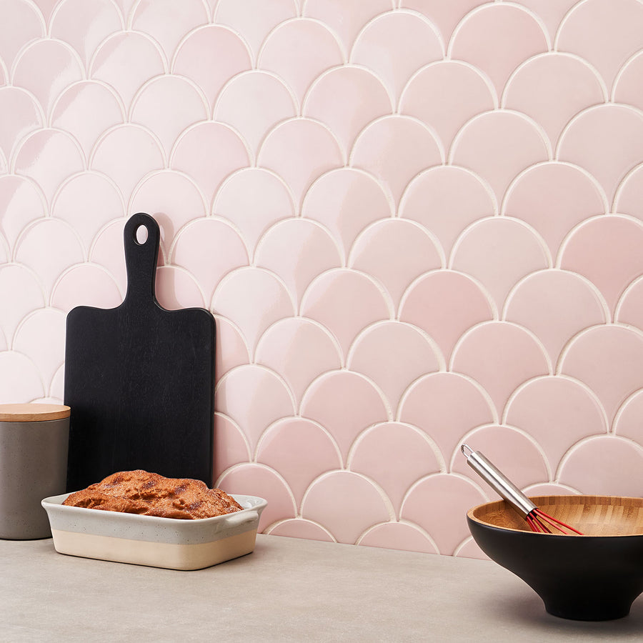 A kitchen counter with a loaf of bread in a baking dish, a black cutting board, a canister, and a wooden bowl with a whisk, set against a pink scallop-patterned tile backsplash.