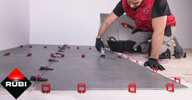 A person wearing gloves and a red vest kneels on the floor installing large gray tiles using red and black Rubi Delta Leveling System spacers; the RUBI logo appears in the lower left corner.