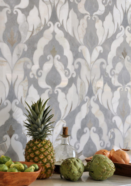 A pineapple, artichokes, onions, and Brussels sprouts sit on a kitchen counter with a glass bottle in front of a gray and white, ornate patterned tile backsplash.