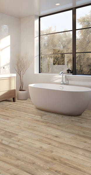 A modern bathroom with a freestanding oval bathtub, large windows showing trees outside, light wood flooring, a white vanity with drawers, and a minimalist potted tree in the corner.