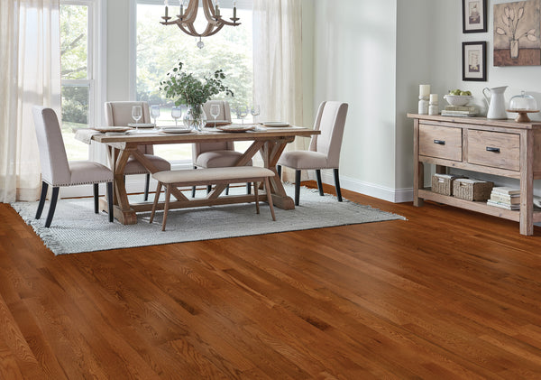 A bright dining room with hardwood floors, a wooden dining table set for four, a bench, chairs, a sideboard, and large windows with sheer curtains letting in natural light.