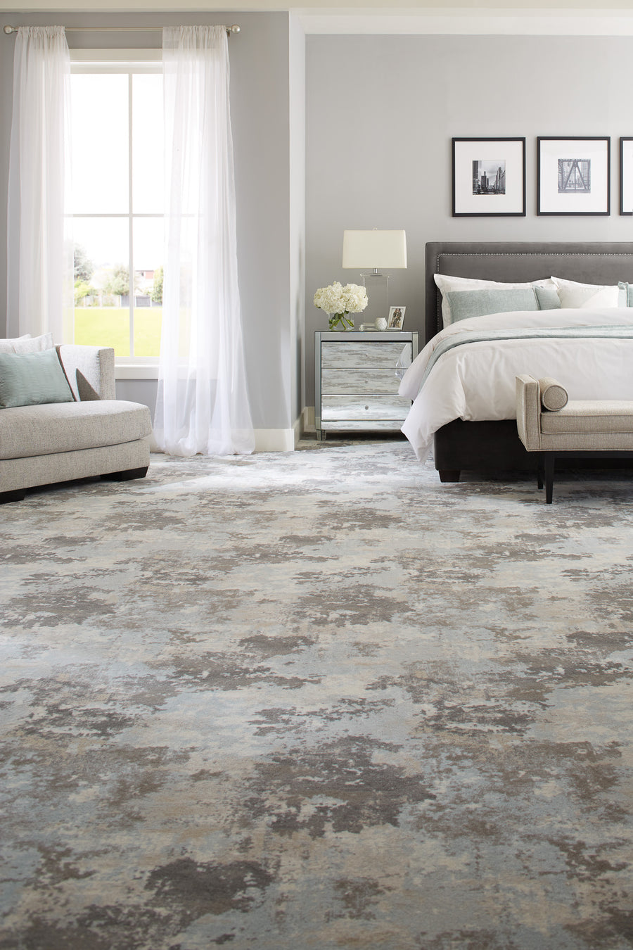 A modern bedroom with a large, abstract-patterned rug, a bed with white bedding, a mirrored nightstand, a gray sofa, and a large window with sheer white curtains letting in natural light.