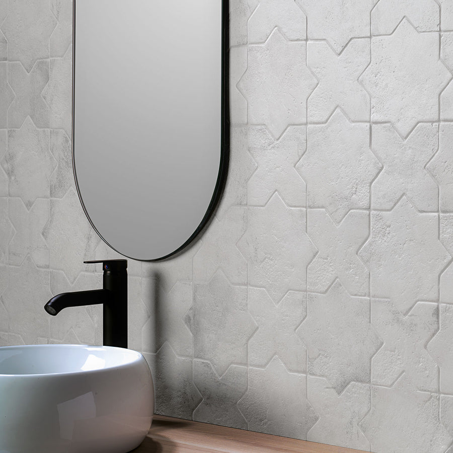 A close-up of a modern bathroom with a white patterned tile wall, a round white vessel sink, a sleek black faucet, and a tall, rounded rectangular mirror above a wooden countertop.