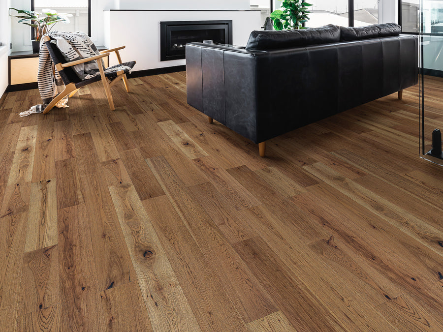 Modern living room with natural wood flooring, a black leather sofa, a wooden lounge chair with a blanket, potted plants, a fireplace, and large windows letting in natural light.