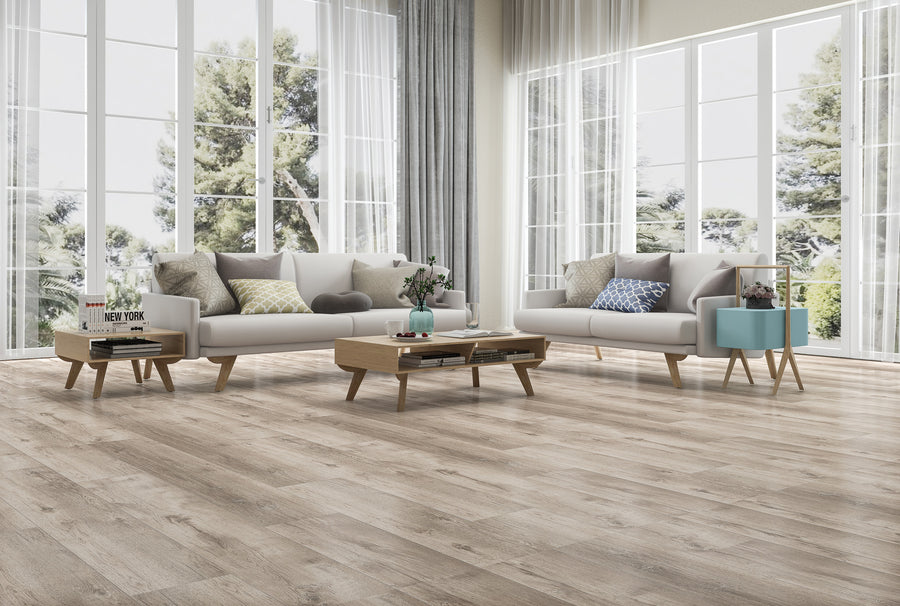 A modern living room with two light sofas, decorative pillows, wooden coffee tables, and floor-to-ceiling windows overlooking trees. The space has light wood flooring and sheer curtains.