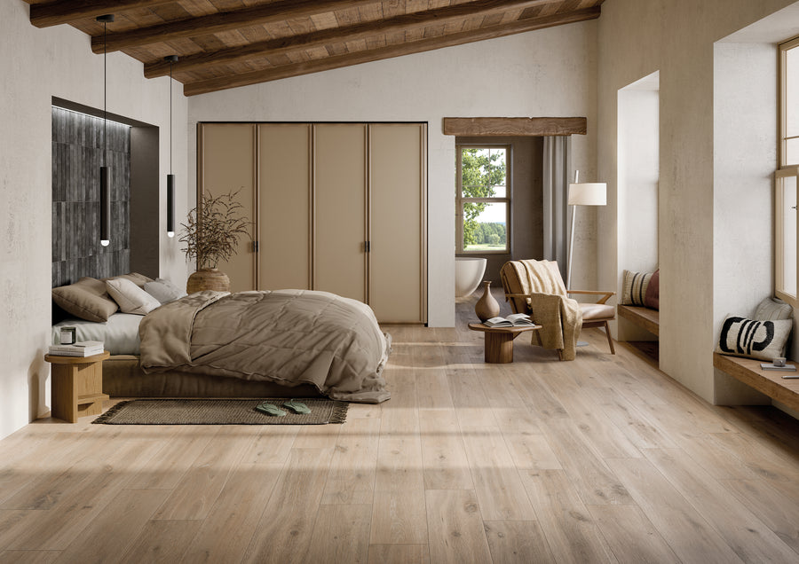 A modern, cozy bedroom with wooden floors, a large bed with beige bedding, built-in tan wardrobes, a lounge chair, a large window seat with cushions, and natural light streaming in from windows.