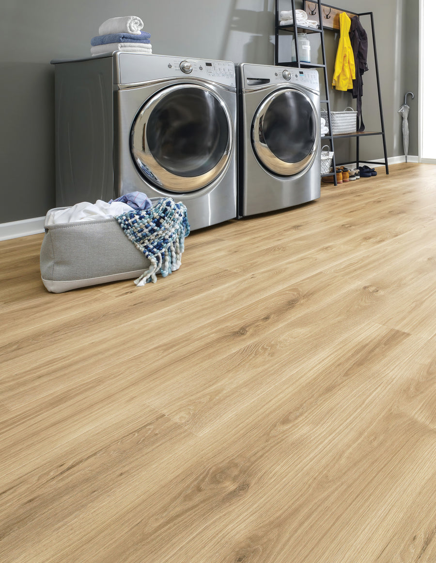 A modern laundry room with a silver washer and dryer, a basket of laundry with a blue and white scarf, shelves with towels, clothes, and accessories, and light wood flooring.