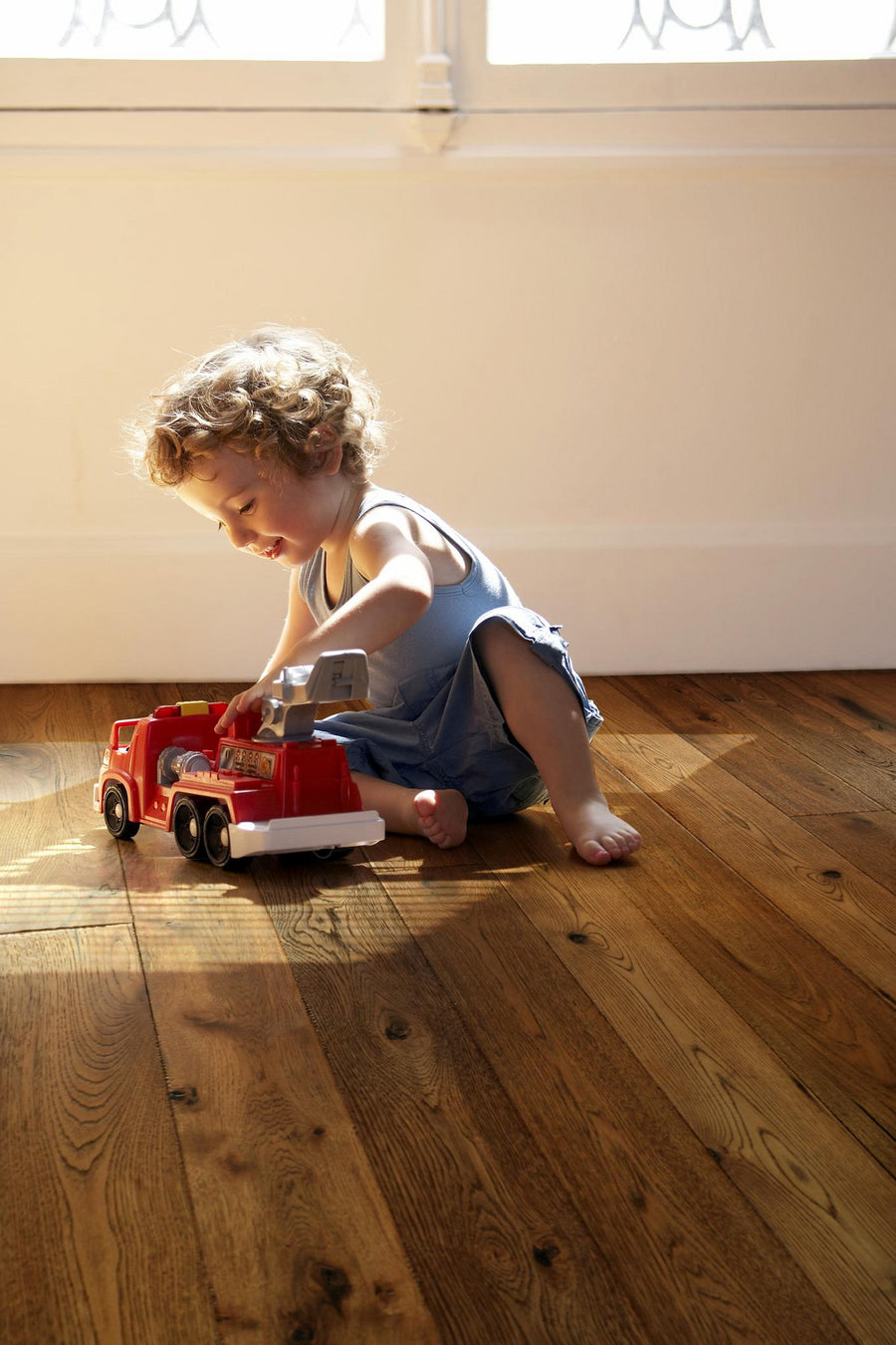 A young child with curly hair sits on a wooden floor, playing with a red toy fire truck in natural sunlight near a window.