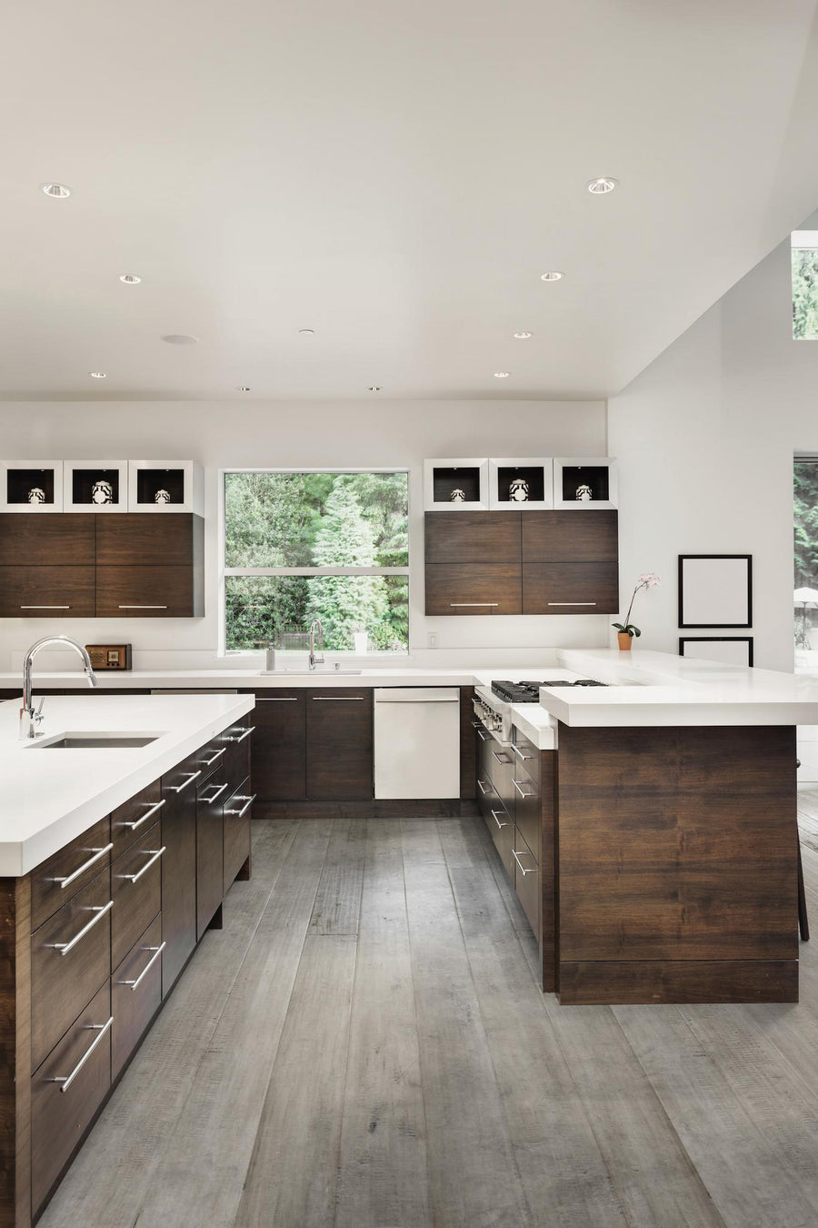 Modern kitchen with dark wood cabinets, white countertops, an island, large windows, light wood floors, and minimalist decor. The space is bright and open with recessed lighting and a view of greenery outside.
