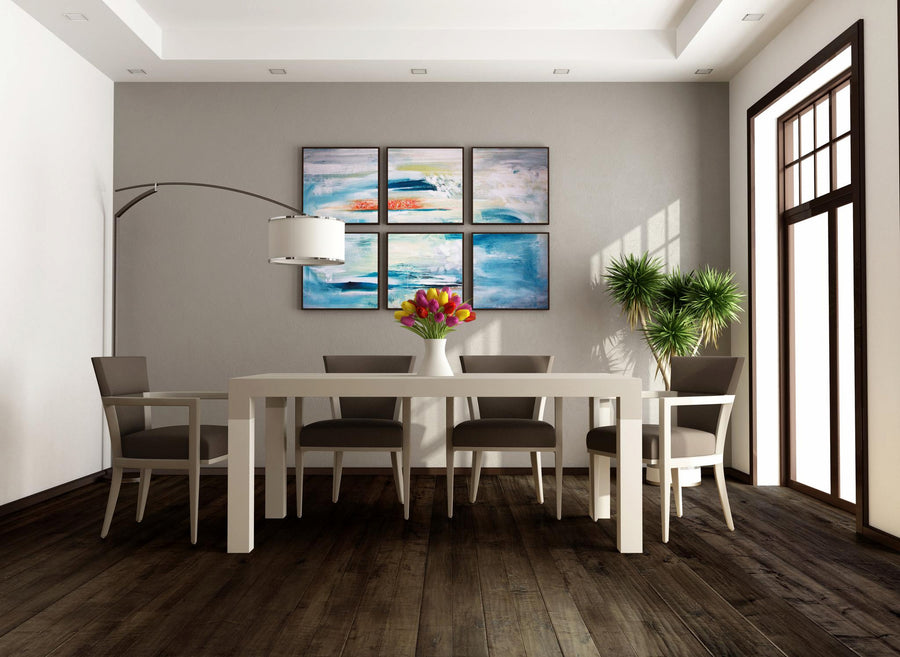 Modern dining room with a white table, six gray chairs, a vase of colorful flowers, indoor potted plant, large window, and four abstract paintings with blue and orange tones on a gray wall.