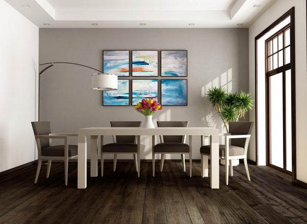 Modern dining room with a white table, six gray chairs, a vase of colorful flowers, indoor potted plant, large window, and four abstract paintings with blue and orange tones on a gray wall.