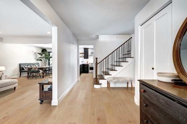 Bright, modern hallway with light wood floors, a staircase with dark railings, a round mirror on a wooden dresser, and a view into a living room and kitchen with plants and dining furniture.