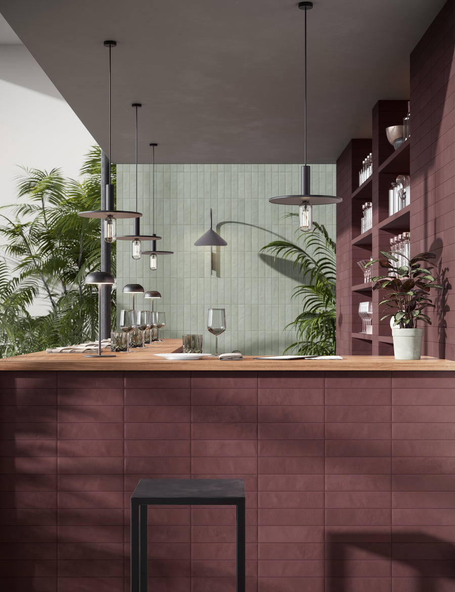 A modern bar area with maroon tile front, a wooden countertop, hanging pendant lights, empty glassware, green plants, and open shelves against pastel green and maroon tiled walls.