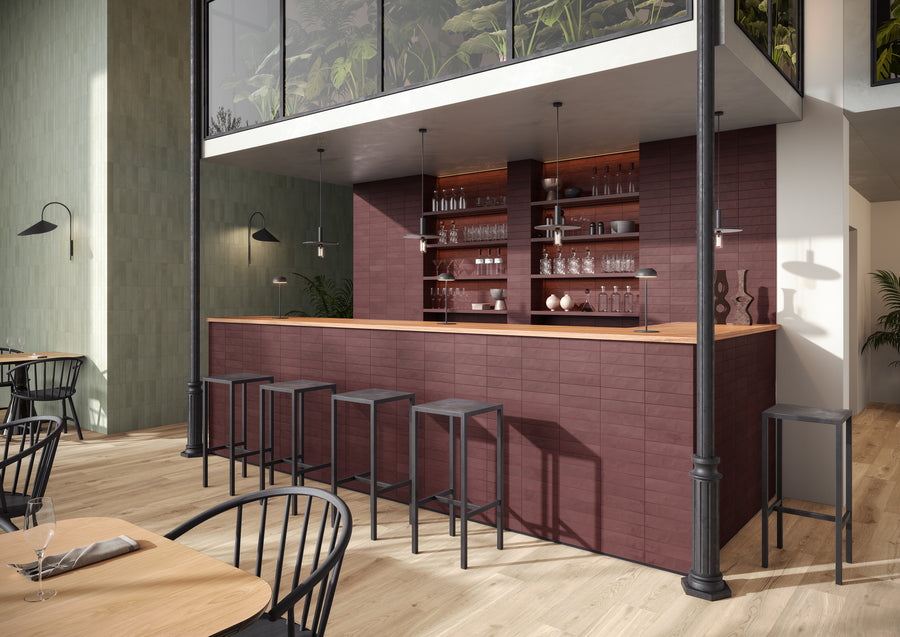 A modern bar area with a burgundy tiled counter, wooden countertop, black bar stools, glass shelves with bottles and glasses, light wood flooring, and green plants visible on an upper level.