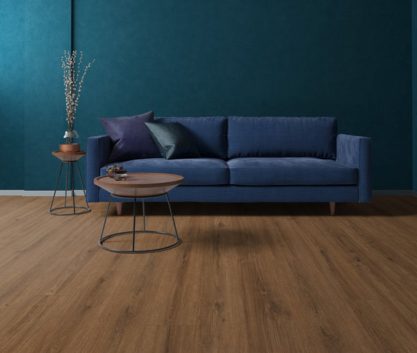 A modern living room with a blue sofa, two round wooden tables, dark teal walls, wood flooring, and a vase with decorative branches on a side table. Pillows are arranged neatly on the sofa.