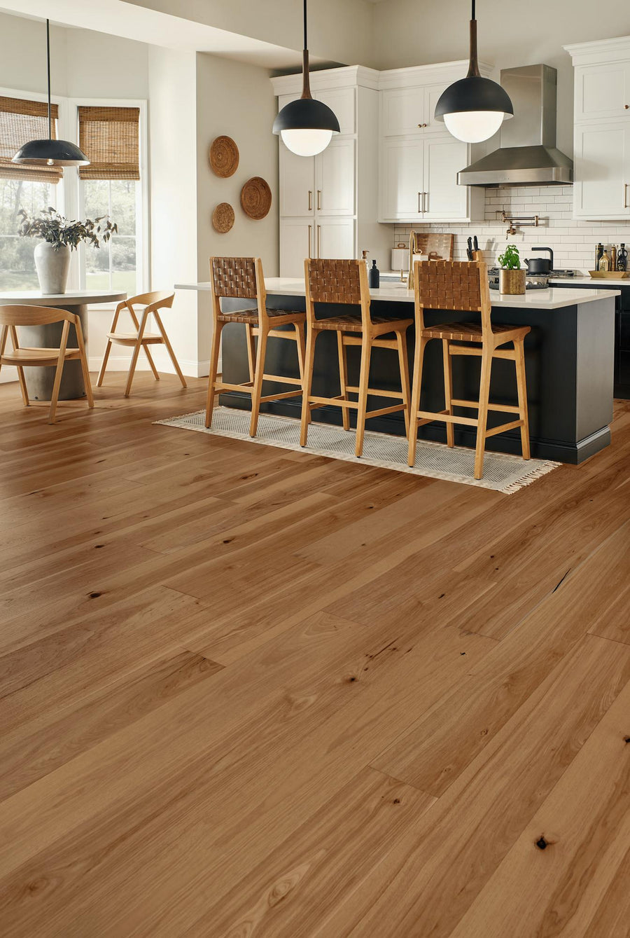 Modern kitchen with light wood flooring, white cabinets, a large dark island with four woven bar stools, pendant lights, and a dining area with wooden chairs near large windows. Natural light fills the space.