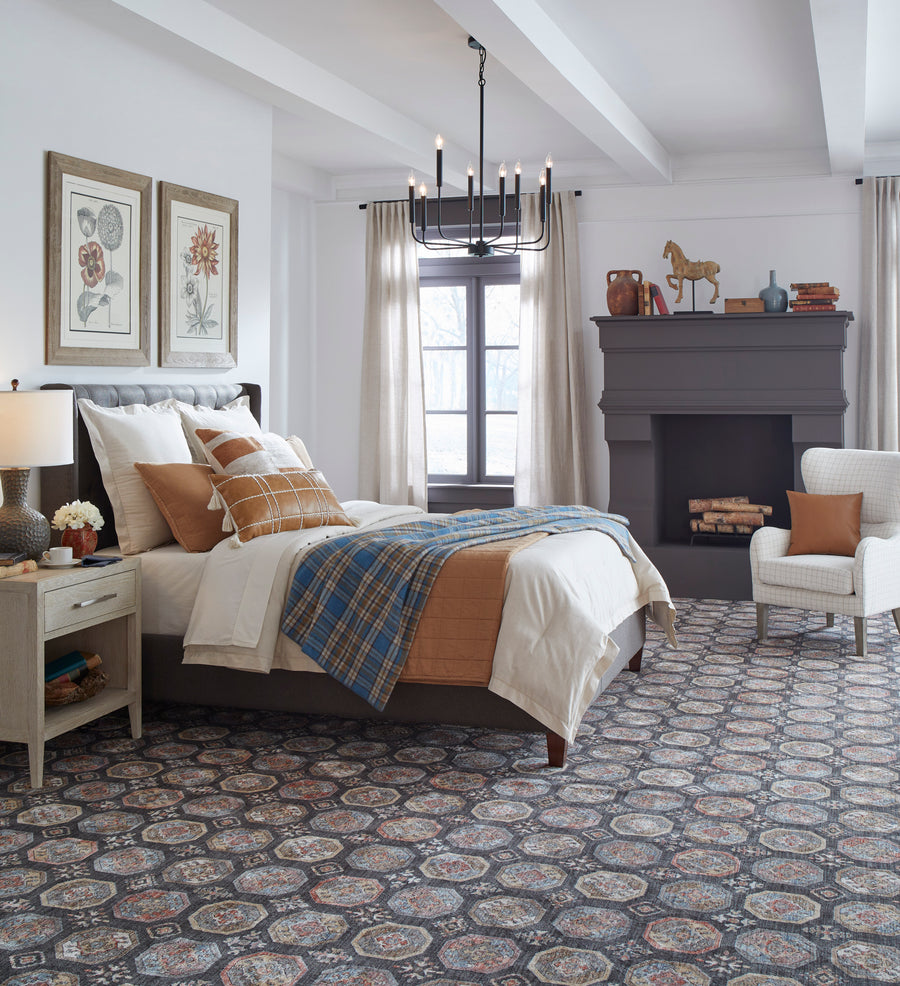 A cozy bedroom with a patterned carpet, neatly made bed with blue and orange bedding, two framed floral prints on the wall, a dark gray fireplace, chandelier, armchair, and large window with light curtains.