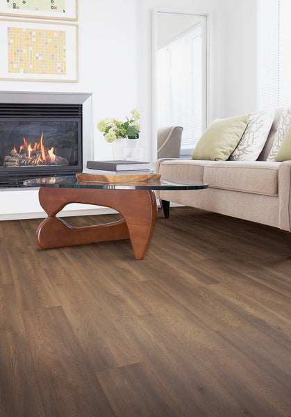 A modern living room with wood flooring, a beige sofa with pillows, a glass coffee table with a curved wooden base, a fireplace with a fire burning, a vase of flowers, and wall art in a bright, airy space.