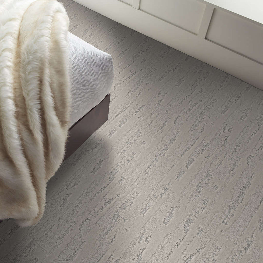 A close-up view of a textured light gray carpet next to a bed with a soft, patterned blanket and a plush cream throw, set against a white paneled wall.