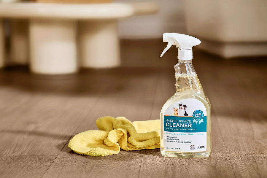 A bottle of Shaw Hard Surface 32OZ Spray Cleaner and a yellow cloth rest on a wooden floor in a home, with a blurred table and chair behind—ideal for keeping hardwood floors spotless.