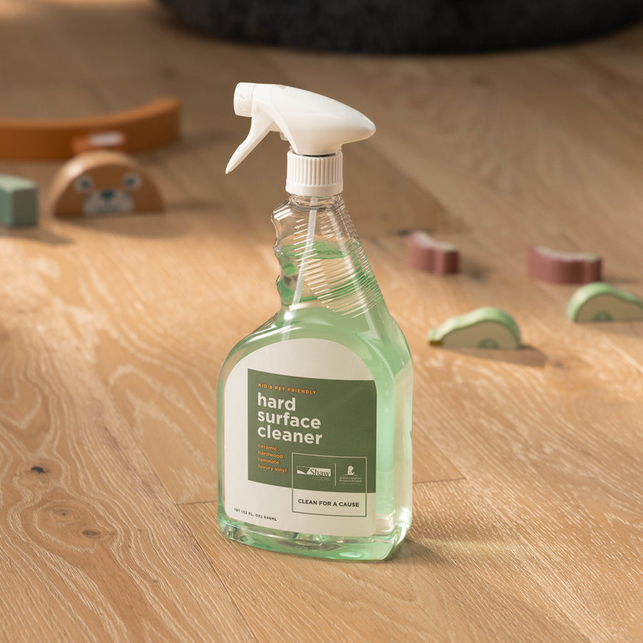 A bottle of Shaw Hard Surface 32OZ Spray Cleaner rests on a wooden floor, while children's wooden toys are scattered in the background.