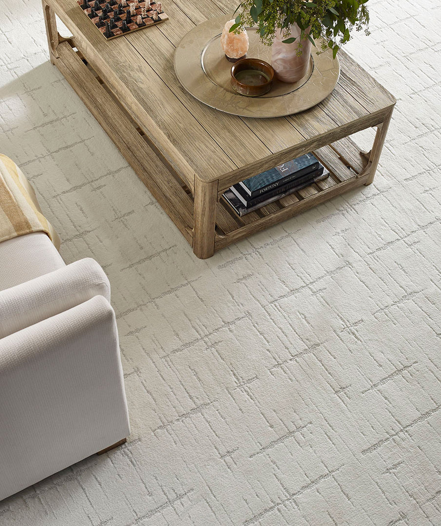 A light-colored, textured rug with a subtle geometric pattern sits beneath a wooden coffee table, which holds books, a tray, a small plant, and decorative items; part of a light sofa is visible on the left.