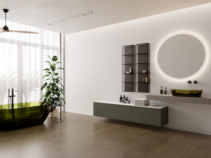 Modern bathroom with a green-tinted glass bathtub, large round backlit mirror above a floating sink, wall shelves with toiletries, and a tall plant by a window with sheer white curtains.