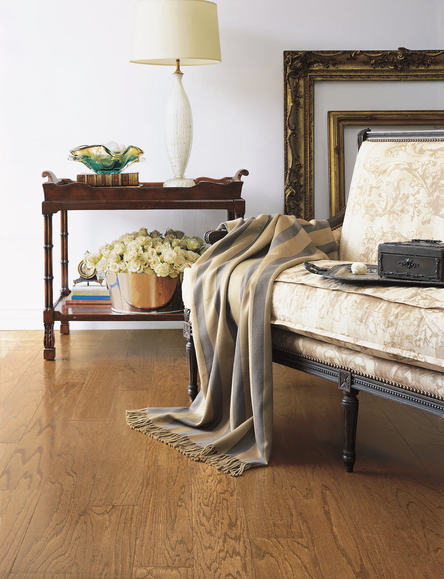 A cozy living room features a vintage sofa with a patterned throw blanket, a side table topped with a lamp and decor, a vase of white flowers, and empty ornate frames leaning against a white wall on a wooden floor.