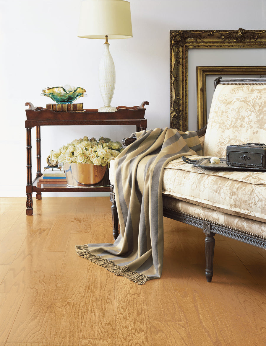 A cozy living room corner with a vintage sofa draped with a striped throw, a side table holding a lamp and decorative items, and a large bowl of white flowers on the floor beside wooden picture frames.