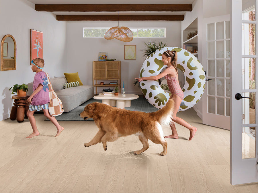 Two children in swimsuits and a golden retriever walk through a bright living room; one child carries a towel and bag, the other carries a large pool float. The room is decorated in a modern, cozy style with natural light.