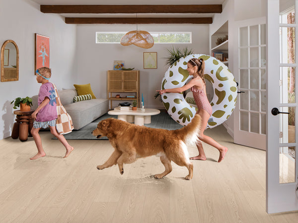 Two children in swimsuits and a golden retriever walk through a bright living room; one child carries a towel and bag, the other carries a large pool float. The room is decorated in a modern, cozy style with natural light.