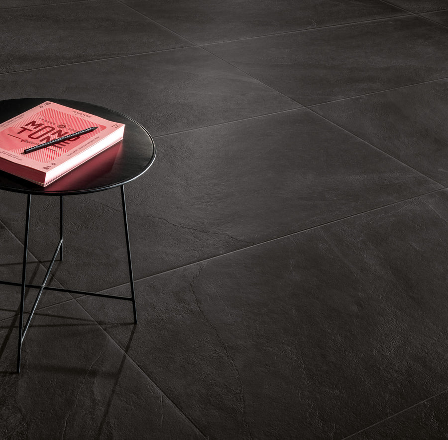 A round, black side table with thin legs holds a pink book. The table stands on large, dark gray textured floor tiles. The scene is minimalistic with ample empty space.
