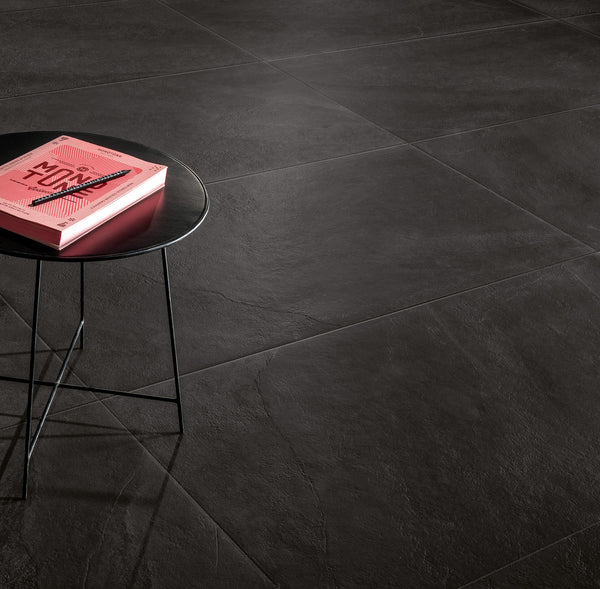 A round, black side table with thin legs holds a pink book. The table stands on large, dark gray textured floor tiles. The scene is minimalistic with ample empty space.