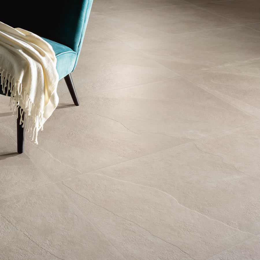 A teal chair with wooden legs and a cream-colored throw sits on large, light beige, textured floor tiles with subtle natural stone patterns.